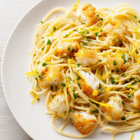 A close-up of Crispy Fish Pasta highlighting the flaky white fish, herb flecks, and glossy lemon sauce in a steaming skillet.