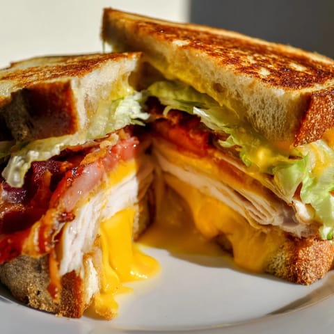 Sizzling slices of Turkey Club Grilled Cheese in a pan, featuring cheddar, bacon, and fresh vegetables on buttery bread.  